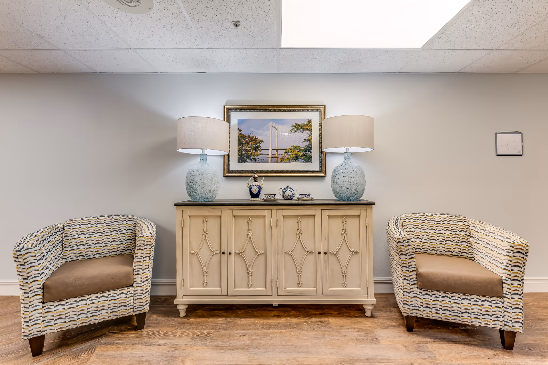 Two patterned armchairs flank a decorative cream cabinet topped with matching lamps and a framed painting in a well-lit seating area.