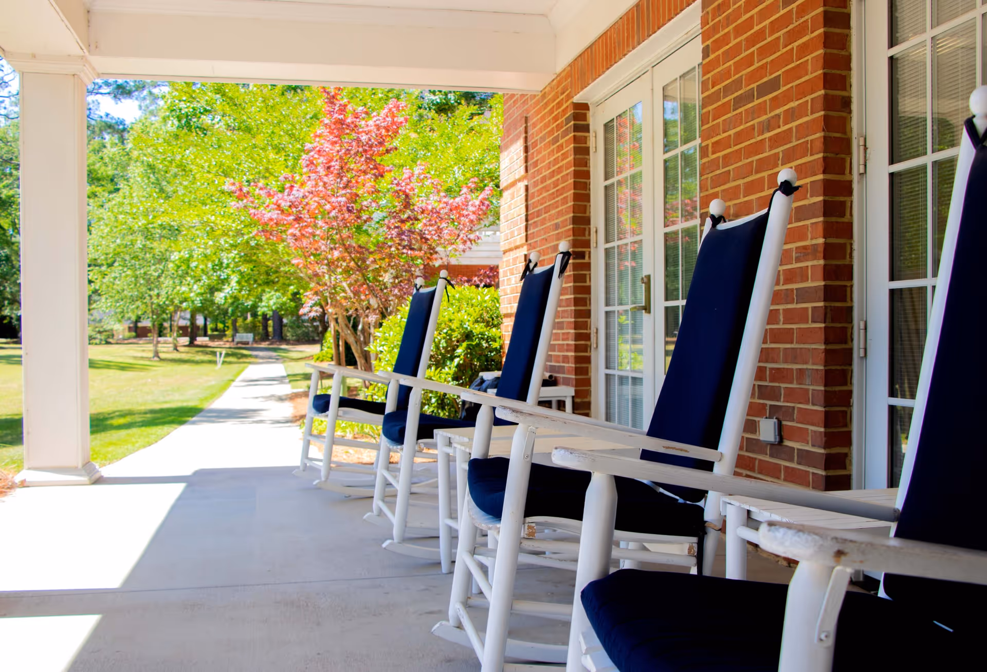 A covered outdoor porch area with white rocking chairs that have dark blue cushions, facing a walkway. The porch is attached to a brick building with large windows and doors. In the background, there are green trees and a small tree with red leaves.