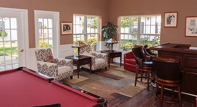 A cozy interior room featuring a red pool table in the foreground, two patterned armchairs with a small table and lamp between them, a corner with a potted plant, and a wooden bar with three bar stools. Large windows and a glass door let in natural light and show greenery outside.