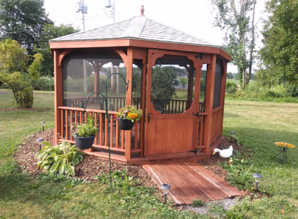 A wooden gazebo with a shingled roof and screened sides sits in a grassy garden area. There are two hanging flower pots with colorful flowers on a metal stand in front of the gazebo. A small white duck figurine is on the ground near the gazebo, and there is a small birdbath to the right. The surrounding area is green with trees and bushes.