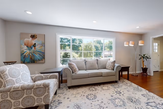 A bright living room with a large window showing greenery outside. The room features a gray sofa with cushions, a patterned armchair, a large area rug with floral designs, two side tables, a floor lamp with three lights, a potted plant, and a painting of a woman in a colorful dress on the wall.