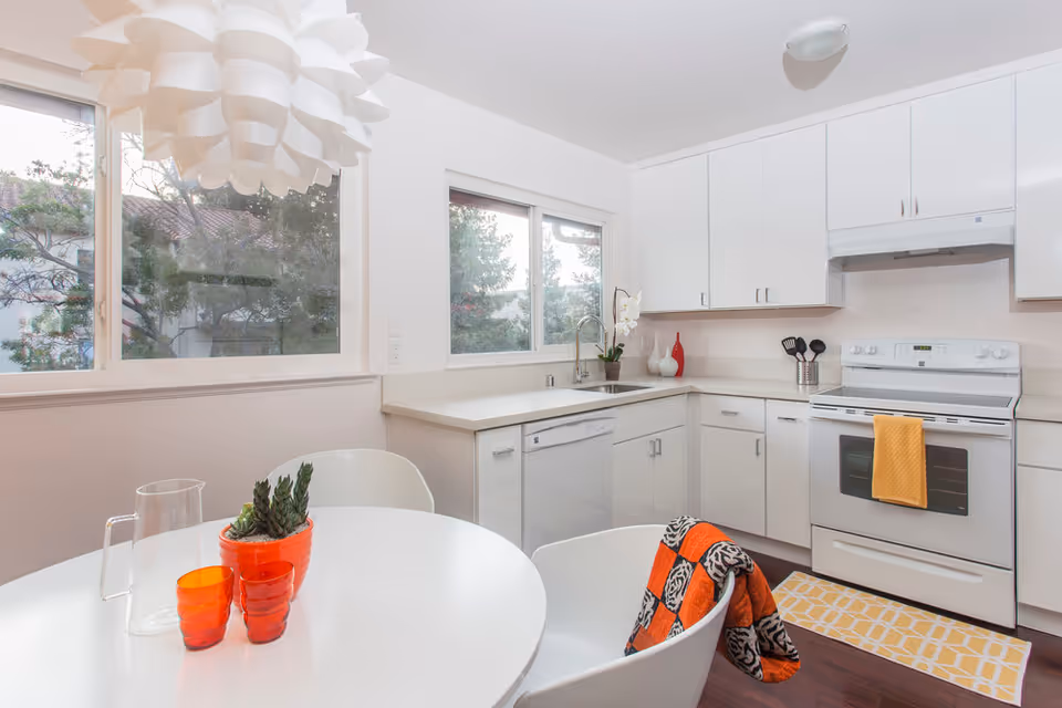 Bright and modern kitchen with white cabinets, a white stove with a yellow towel hanging on the handle, a dishwasher, and a sink under two large windows. A round white table with two white chairs is in the foreground, one chair draped with an orange and black patterned blanket. On the table are two orange glasses, a clear pitcher, and a small potted succulent. A large white pendant light hangs above the table.