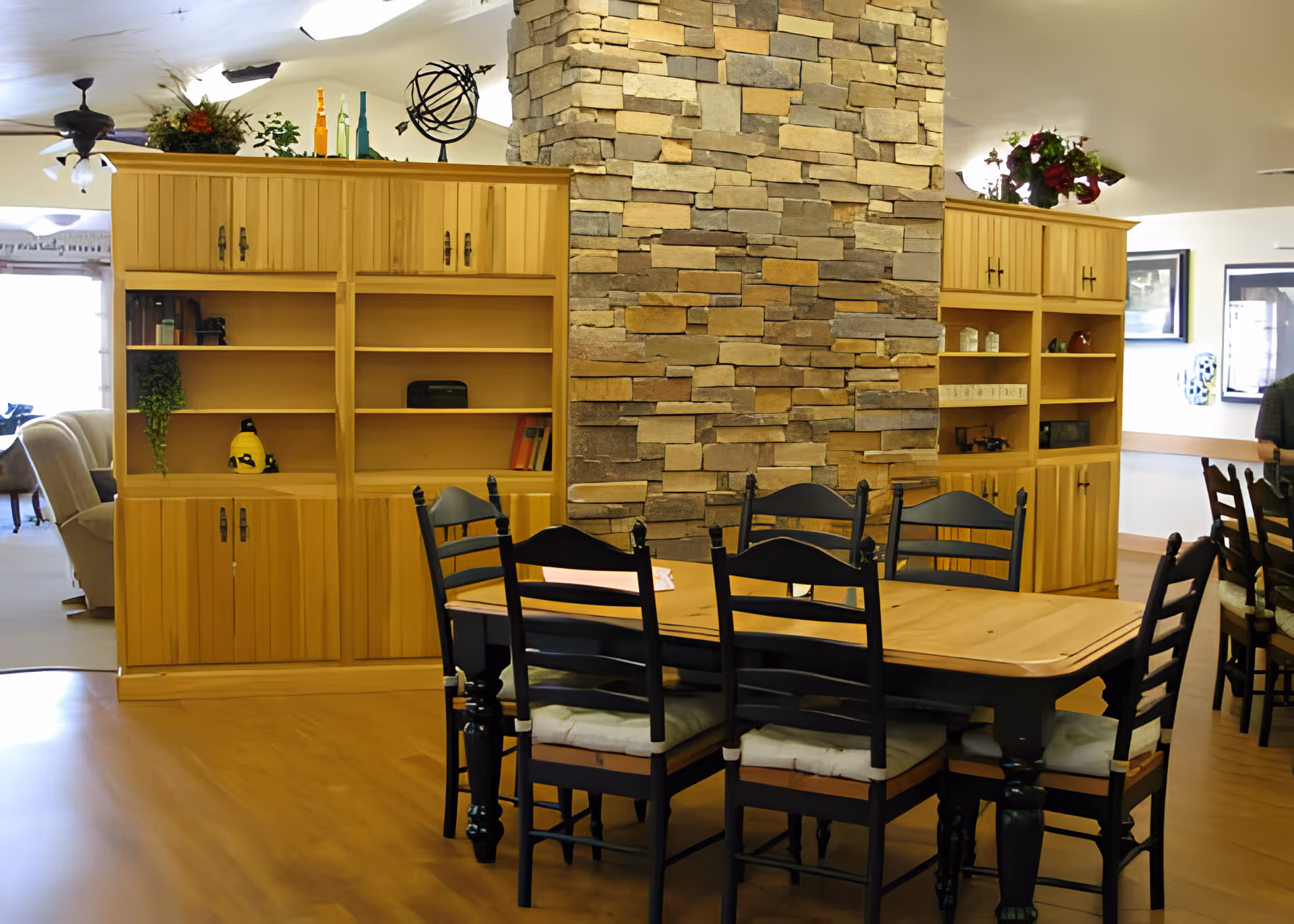 A dining area with a wooden table surrounded by six black chairs with cushions. Behind the table is a stone fireplace and two wooden shelving units with cabinets, decorated with plants, books, and decorative items. The room has a warm, cozy atmosphere with wooden flooring and ceiling fans.