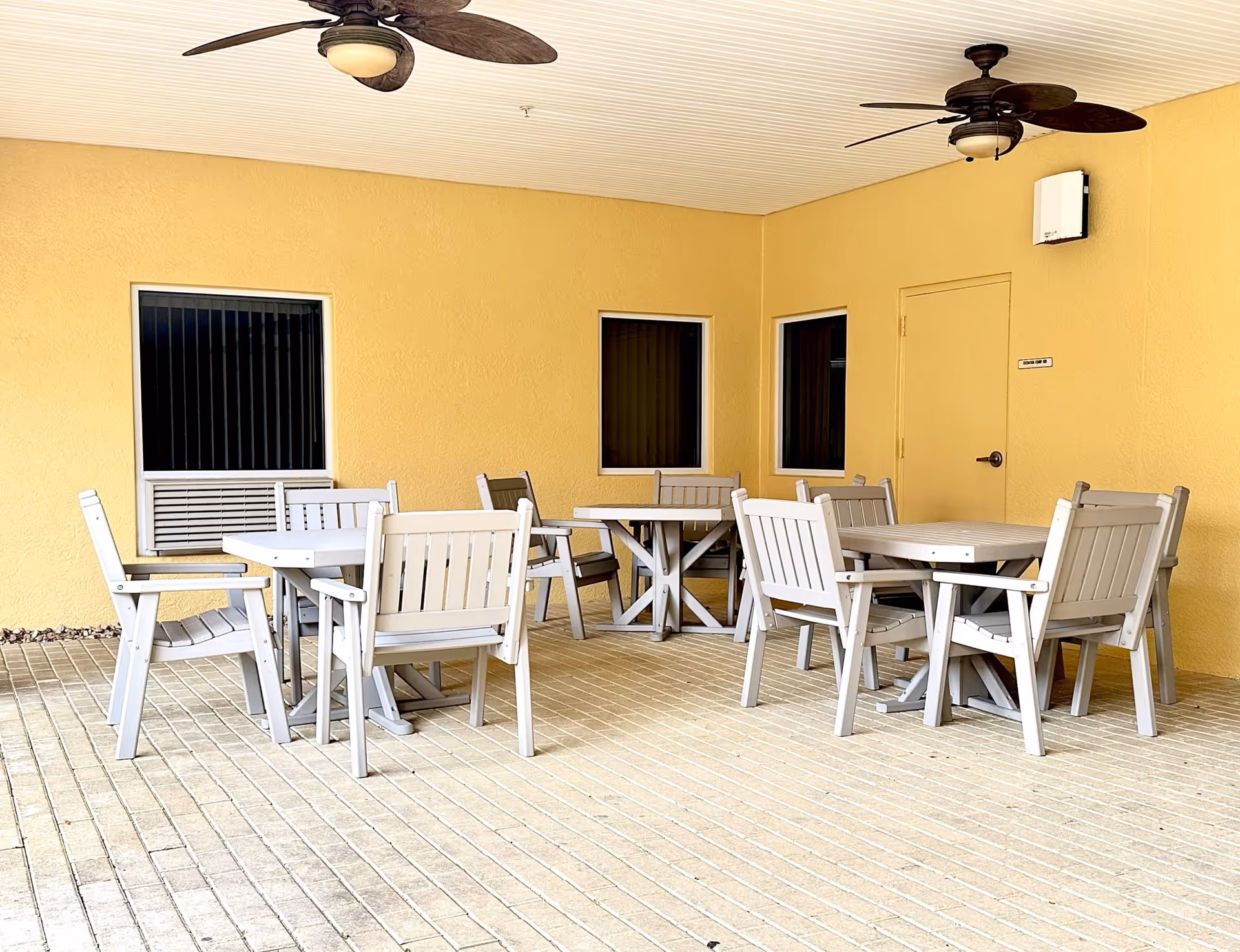 Covered outdoor patio area with yellow walls, two ceiling fans, and three white tables each surrounded by white wooden chairs. There are three windows and a door on the wall.