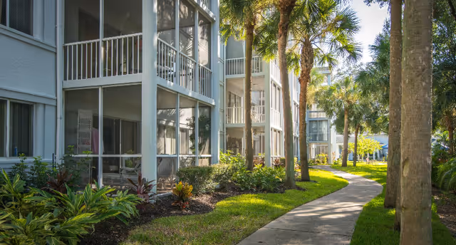 Curved sidewalk with palm trees and landscaping in front of a multi-story senior living building with screened balconies.