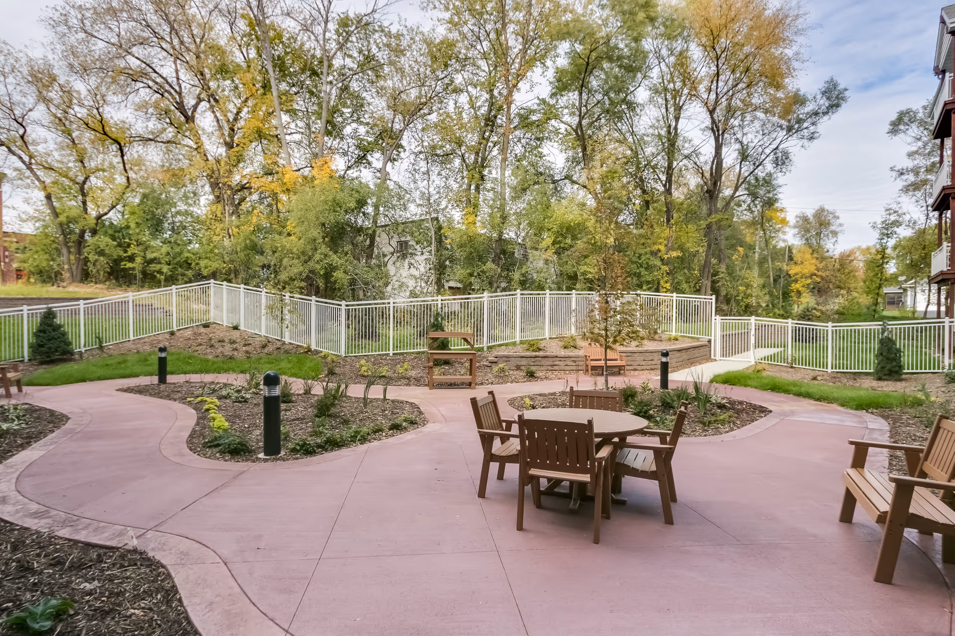 Outdoor courtyard with a circular patio featuring a round table and chairs, benches, curved walkways, landscaped beds and trees behind a white fence.