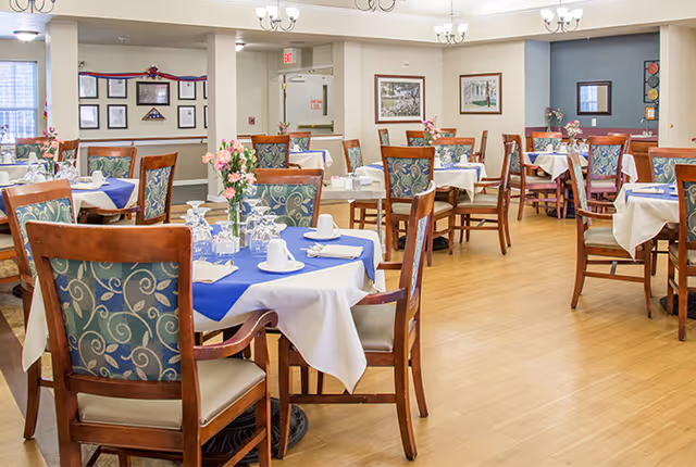 Bright dining room with multiple tables set with white and blue linens, wooden chairs, and floral centerpieces.