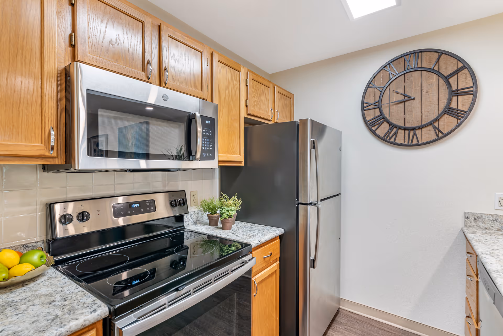 Modern kitchen with wooden cabinets, stainless steel microwave and refrigerator, electric stove with a glass cooktop, granite countertops, two small potted plants, a bowl of lemons and apples, and a large rustic wall clock with Roman numerals on a white wall.
