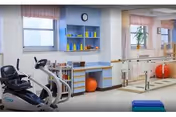 A bright physical therapy or rehabilitation room with exercise equipment including a recumbent bike, parallel bars for walking practice, therapy balls, and shelves with various therapy tools. The room has large windows with pink valances and light blue walls.