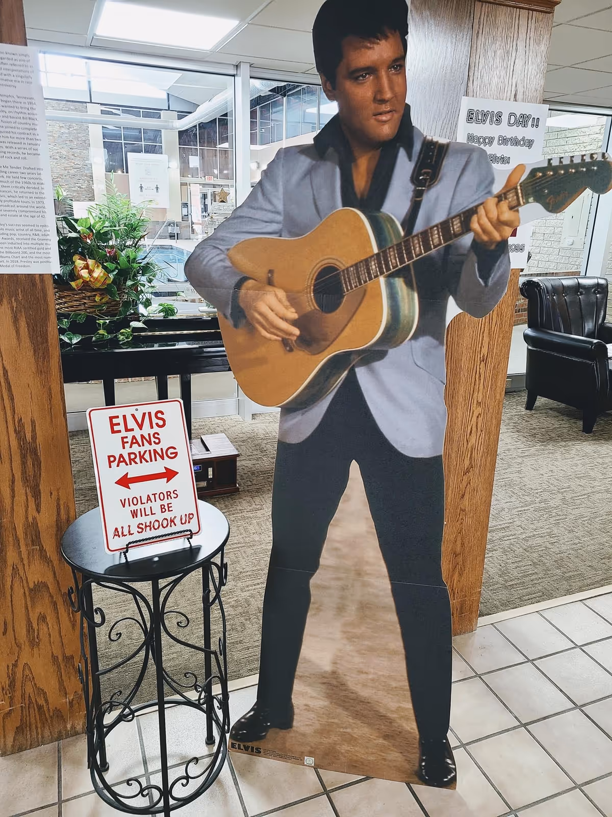 Cardboard cutout of Elvis holding a guitar in a lobby/common area next to a small table with an 'Elvis Fans Parking' sign.