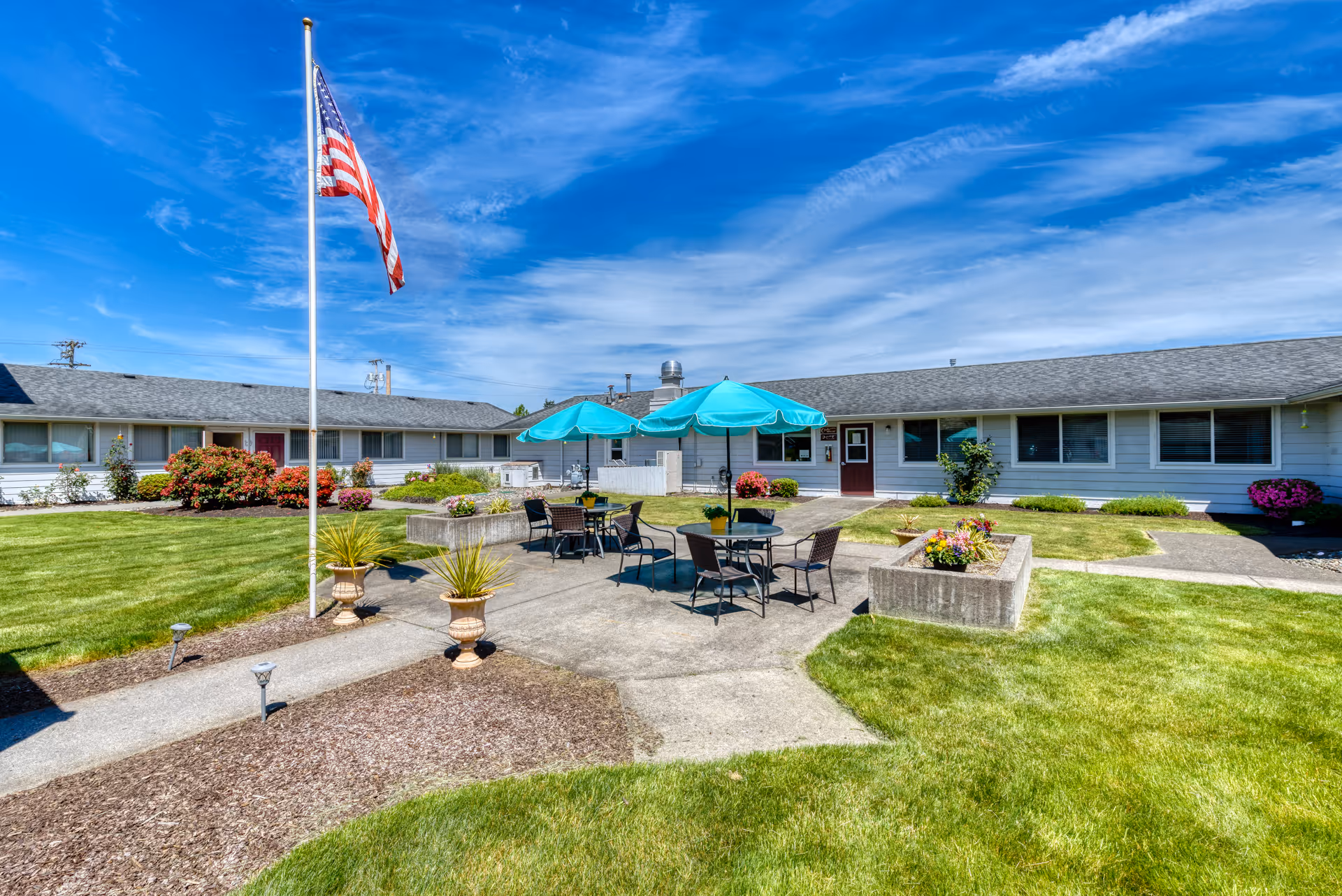 Outdoor courtyard area of Beehive Retirement Community with green grass, flower beds, concrete walkways, tables with turquoise umbrellas, and an American flag on a flagpole under a blue sky with wispy clouds.