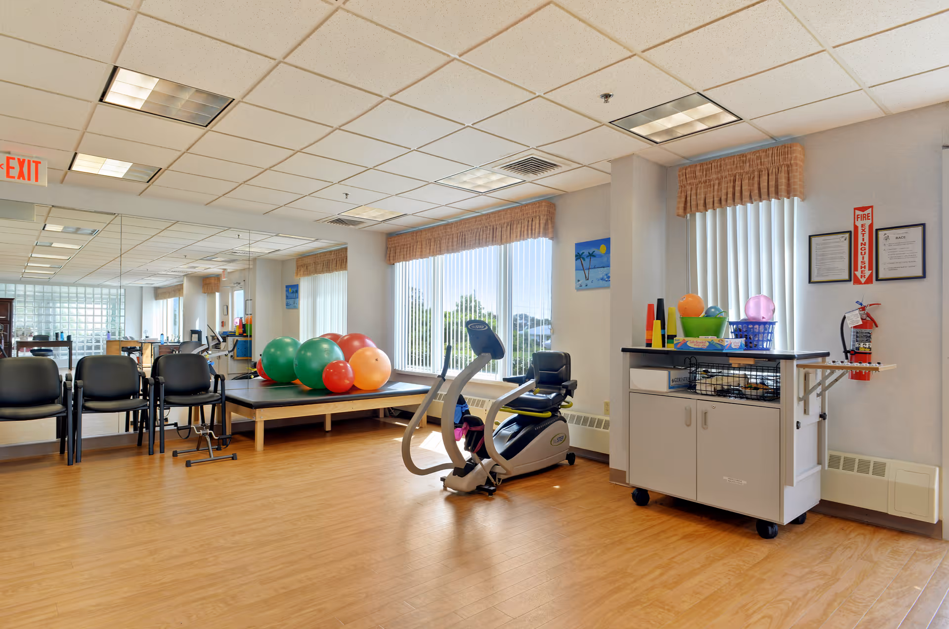 Bright therapy/exercise room with a recumbent exercise bike, treatment table stacked with colorful exercise balls, chairs, and a storage cart.