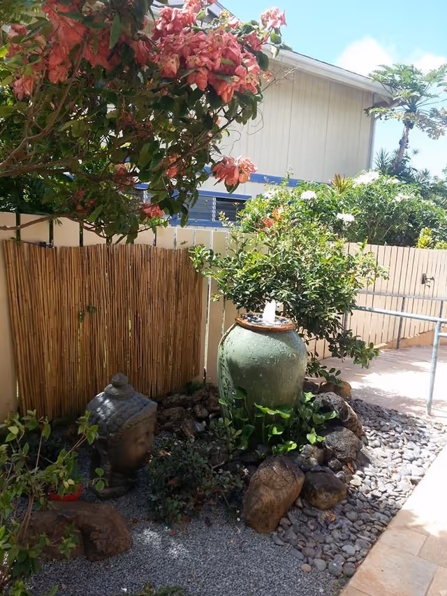 Outdoor garden area with a large green ceramic water fountain surrounded by rocks and plants, a Buddha head statue partially buried in gravel, a bamboo fence, and flowering bushes under a clear blue sky.