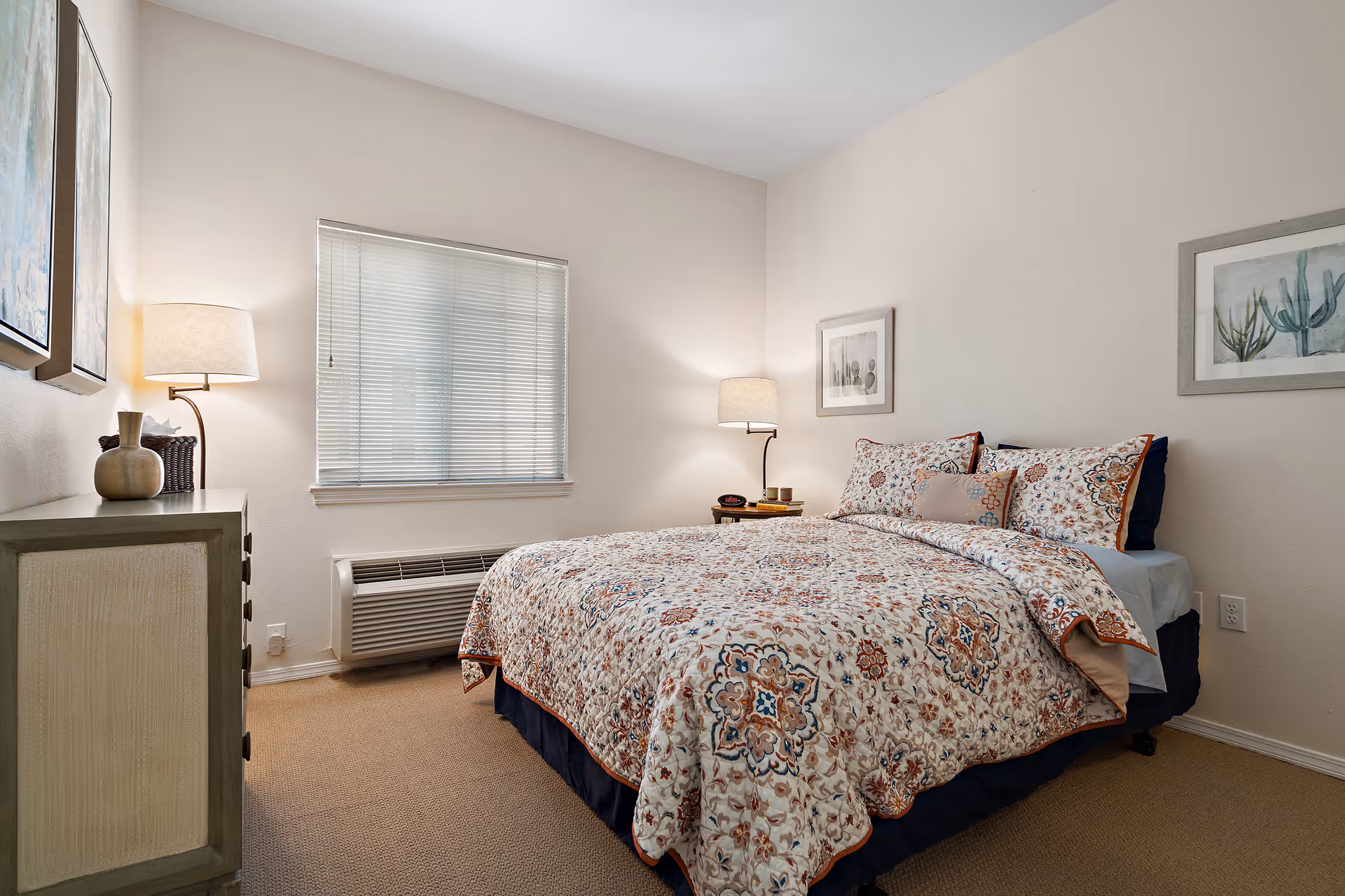 A cozy bedroom with a bed covered in a patterned quilt and matching pillows. There are two framed pictures on the walls, a window with blinds, a dresser with decorative items, and two lamps providing warm lighting.