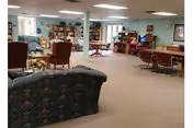 A spacious common area in a retirement facility with various seating options including armchairs and sofas arranged around the room. There are bookshelves, tables, and a television visible, creating a comfortable and inviting living room atmosphere.