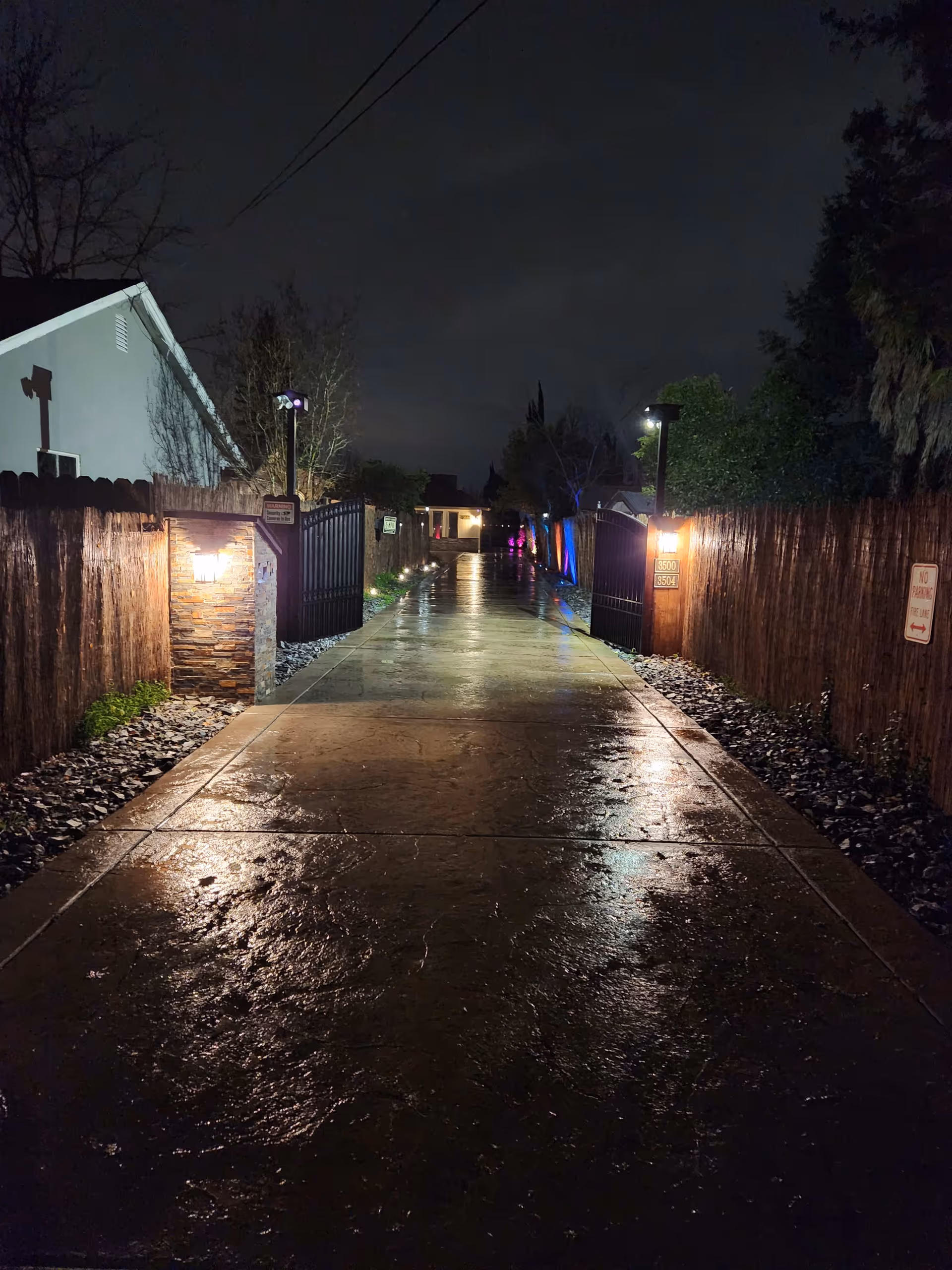 A wet, illuminated pathway at night leading through an open gate flanked by wooden fences and stone pillars with lights. Trees and bushes line the path, and a house is visible in the distance under a dark, cloudy sky.