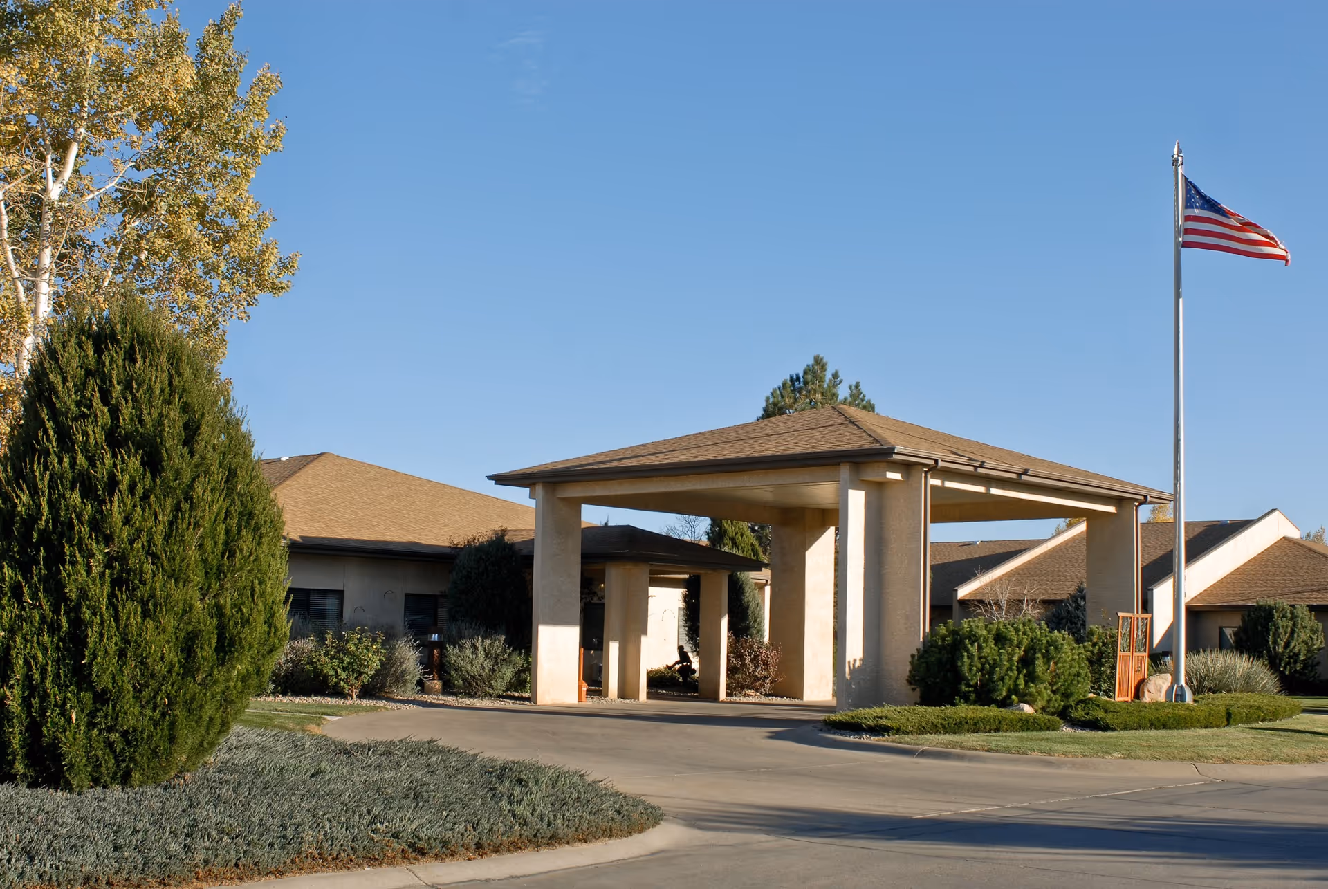 Exterior view of Life Care Center of Pueblo showing the entrance with a covered driveway, surrounding greenery, and an American flag on a flagpole against a clear blue sky.