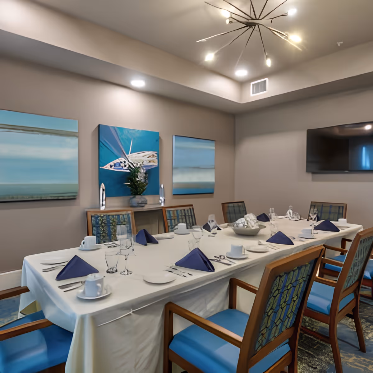 A dining room with a long rectangular table set for a meal, featuring white tablecloth, blue folded napkins, white plates, cups, and glassware. The room has wooden chairs with blue cushions and patterned backs. Three abstract paintings hang on the beige wall, and a modern chandelier with multiple light bulbs is mounted on the ceiling. A flat-screen TV is mounted on the wall in the background.