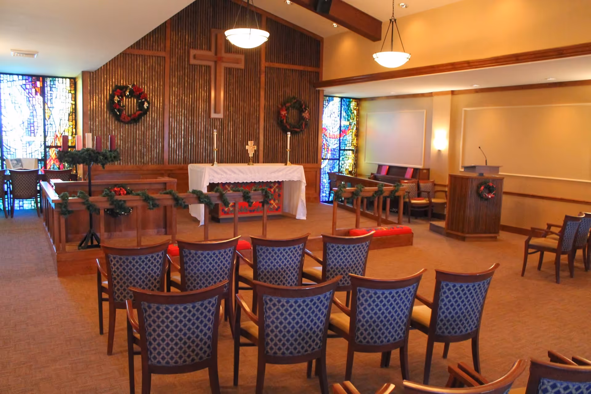 Interior of a chapel or worship room with wooden chairs arranged in rows facing an altar. The altar is decorated with a white cloth, candles, and a cross mounted on a wooden paneled wall. There are stained glass windows on either side of the altar and Christmas wreaths and garlands decorating the space.