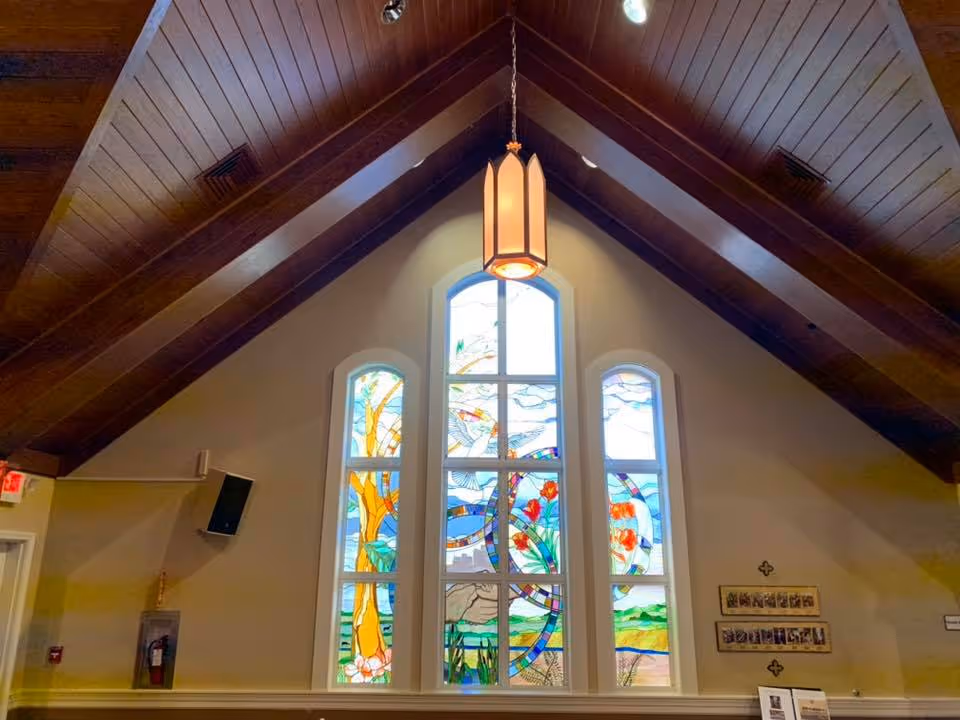Vaulted wood-ceilinged interior with a hanging lantern light and three tall stained-glass windows.