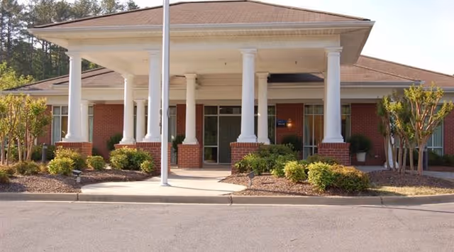 Front exterior view of a single-story brick building with white columns supporting a covered entrance. There are shrubs and small trees planted around the entrance area, and a paved driveway in front.