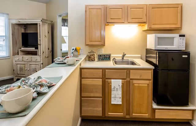 Small kitchen area with light wood cabinets, a stainless steel sink, a black mini refrigerator with a white microwave on top, and a countertop with a decorative sign and a towel hanging from a cabinet. Adjacent to the kitchen is a counter with bowls and cloth napkins, and in the background, a wooden cabinet with shelves and drawers is visible near a window.