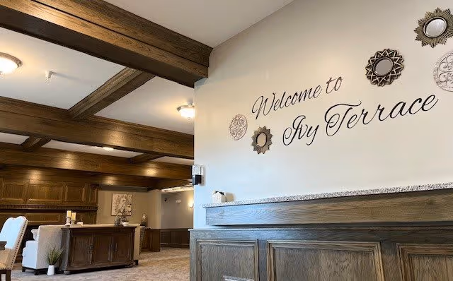 Interior view of a senior living facility with wooden beams on the ceiling, a wooden reception desk, and a wall sign that reads 'Welcome to Ivy Terrace' decorated with ornamental mirrors.