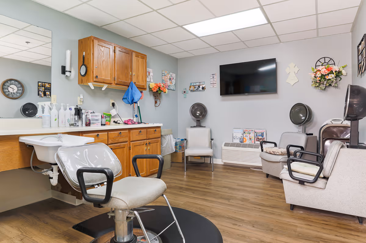 Interior of a salon room in an assisted living facility with a salon chair in front of a sink and counter with various hair care products. The room has wooden cabinets, a large mirror, two hair dryers, a wall-mounted TV, and chairs for waiting. The walls are decorated with flowers, a clock, and family-themed wall art.