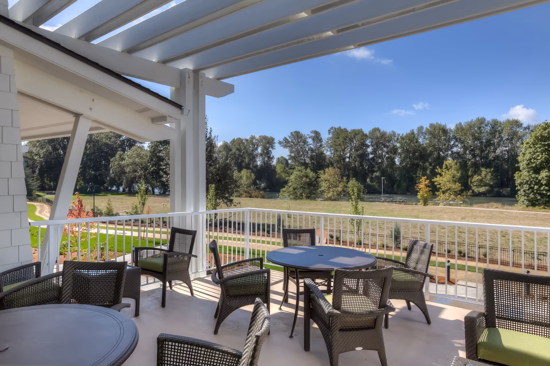Outdoor covered patio area with multiple round tables and wicker chairs with green cushions, overlooking a grassy area with trees and a clear blue sky.