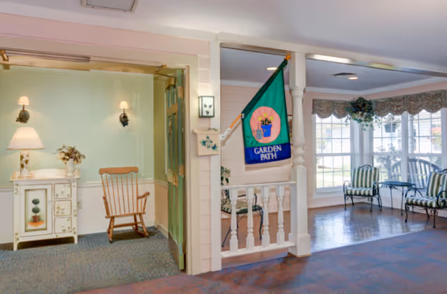 Bright senior living common area with a rocking chair and side table on the left, a decorative 'Garden Path' flag on a white railing, and windowed seating with striped chairs on the right.