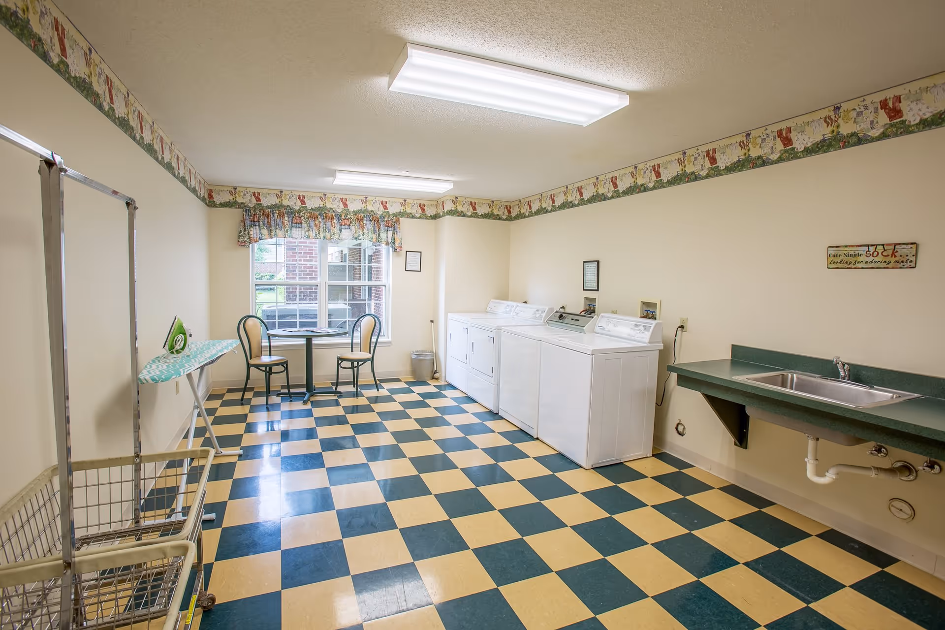 A laundry room with a checkered yellow and green floor, three white washing machines and dryers along one wall, a green countertop with a sink, an ironing board with an iron, a laundry cart, and a small round table with two chairs near a window with floral curtains. The walls are light-colored with a decorative border near the ceiling.