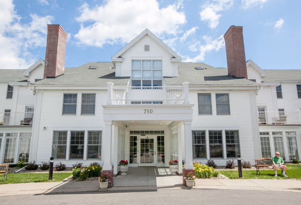 Front exterior of a white multi-story senior living building with an entrance marked "750" and a person sitting on a bench.