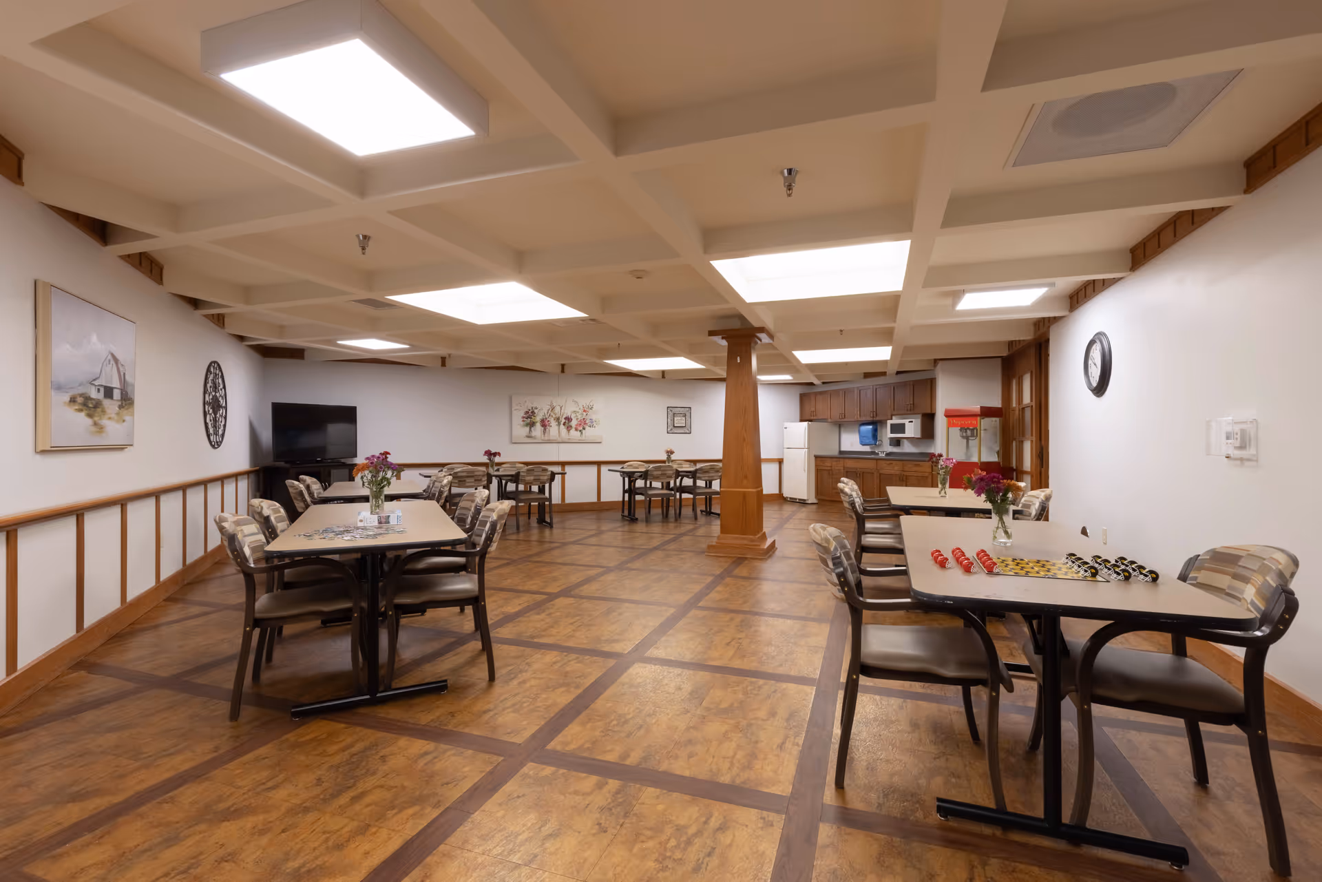 Spacious communal dining/activity room with multiple tables and chairs, a kitchenette, TV, and wall artwork.