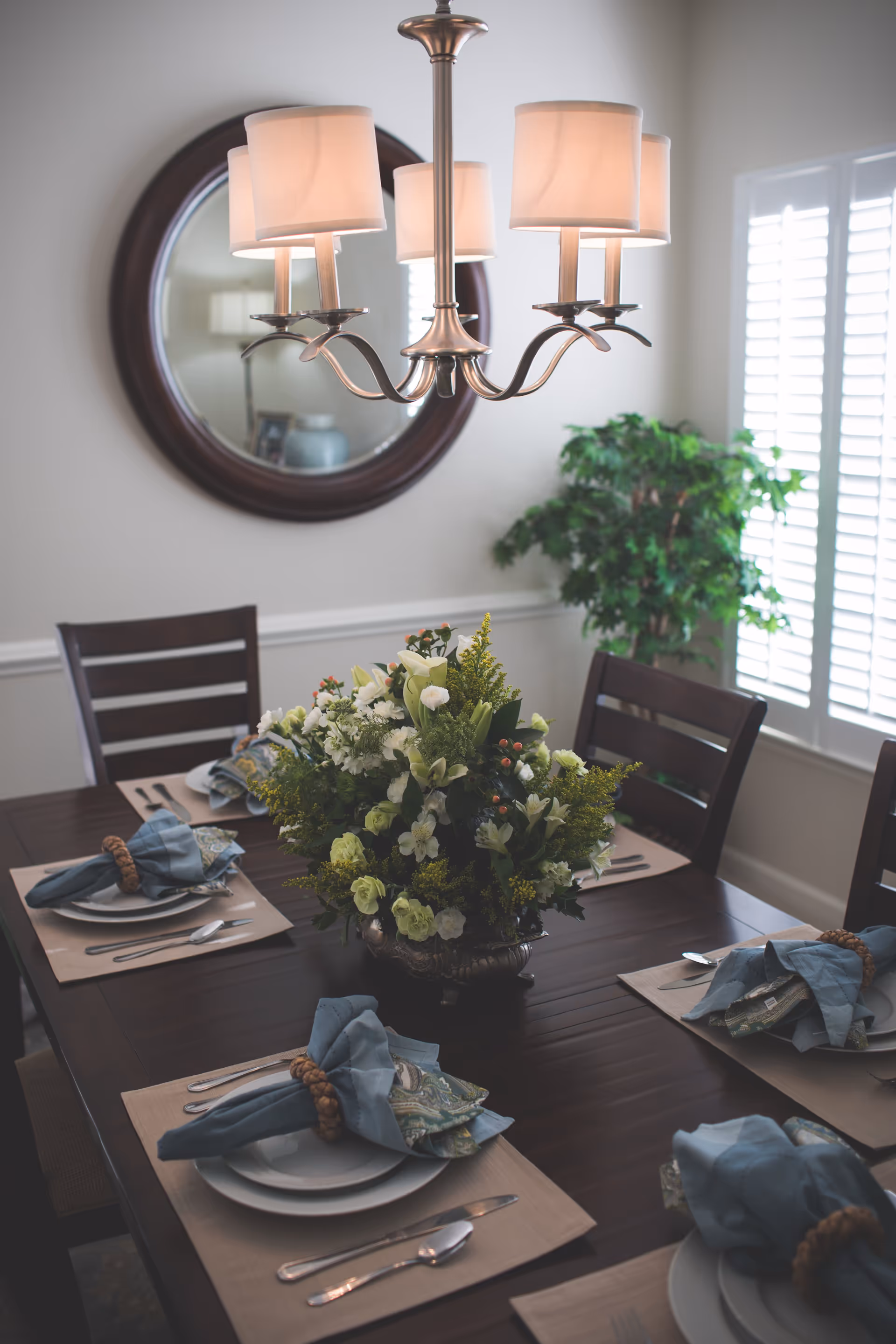 A dining room with a dark wooden table set for six people. Each place setting includes a white plate, silverware, and a blue cloth napkin with a napkin ring. A large floral centerpiece with white and green flowers is in the middle of the table. Above the table hangs a chandelier with five lampshades. In the background, there is a round mirror on the wall and a green potted plant near a window with white shutters.