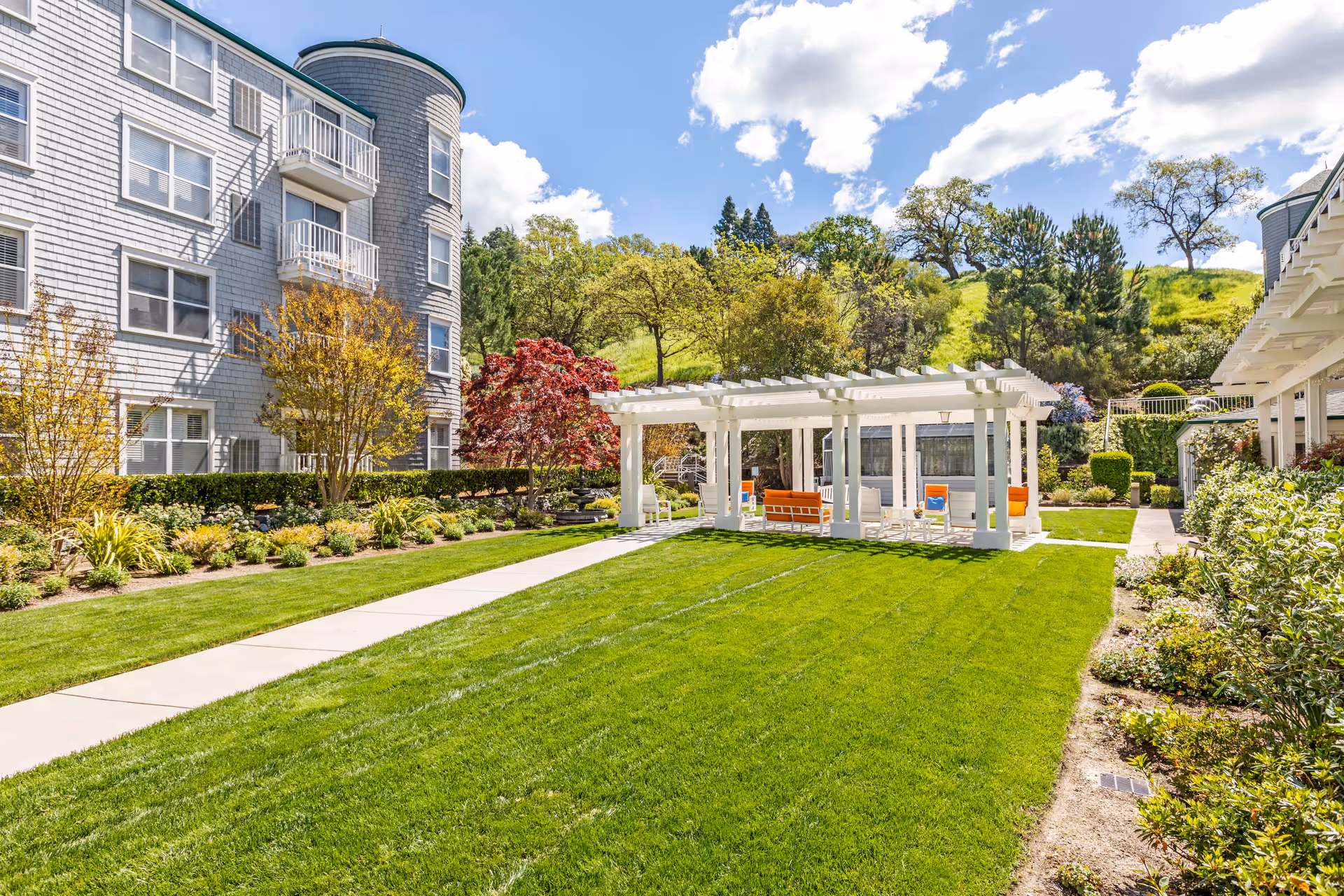 A well-maintained outdoor garden area at Byron Park featuring a green lawn, a white pergola with orange and blue seating underneath, surrounded by trees, shrubs, and a multi-story residential building on the left side under a partly cloudy blue sky.