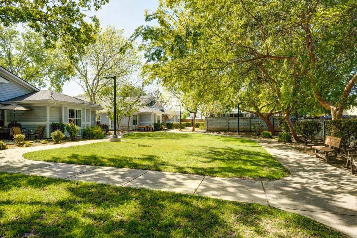 A sunny outdoor courtyard area at a senior living facility with green grass, trees providing shade, paved walkways, benches, and single-story buildings with patios and outdoor seating.