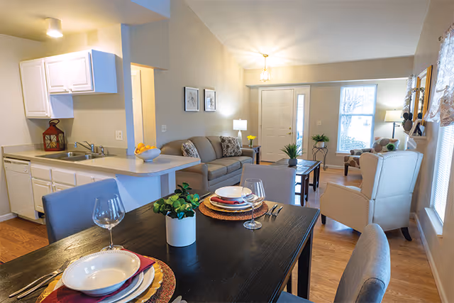 Open-plan interior showing a dining table set for two in the foreground, a kitchenette to the left, and a living area with a sofa and armchairs in the background.