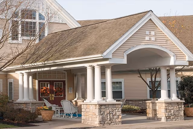 Entrance of a senior living facility with a covered porch supported by white columns and stone bases. There are two red doors decorated with autumn wreaths, outdoor chairs, and potted plants near the entrance. The building has beige siding and a sloped roof.