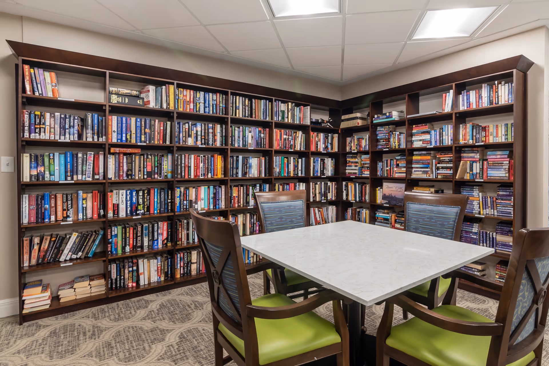 A cozy reading room with dark wooden bookshelves filled with numerous books lining the walls. In the center, there is a square white table surrounded by four wooden chairs with green cushions and patterned backs. The room has a carpeted floor with a subtle pattern and a white drop ceiling.