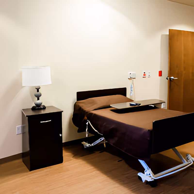 A simple room with a single hospital-style bed covered with a brown blanket, a small bedside table with a lamp, and a wooden door in the background. The room has light-colored walls and a wooden floor.
