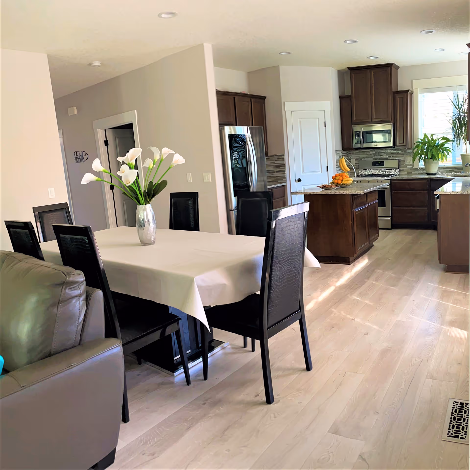 A bright and modern open-plan kitchen and dining area. The dining table is covered with a white tablecloth and has a vase with white flowers as a centerpiece. The kitchen features dark wood cabinets, stainless steel appliances including a refrigerator, stove, and microwave, and a kitchen island with a bowl of fruit. Light wood flooring and neutral-colored walls complete the space.