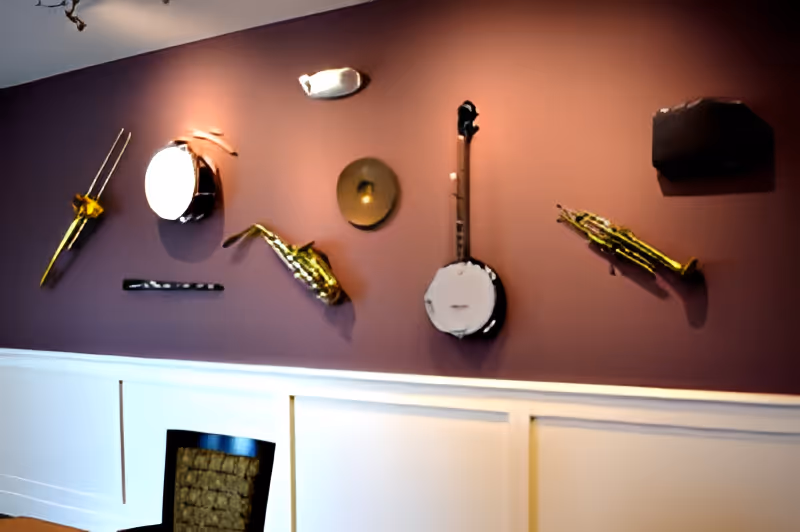 Interior wall painted in a muted purple color with various musical instruments mounted on it, including a trombone, drum, flute, saxophone, cymbal, banjo, and trumpet. Below the instruments is a white wainscoting panel and a chair with a woven backrest.