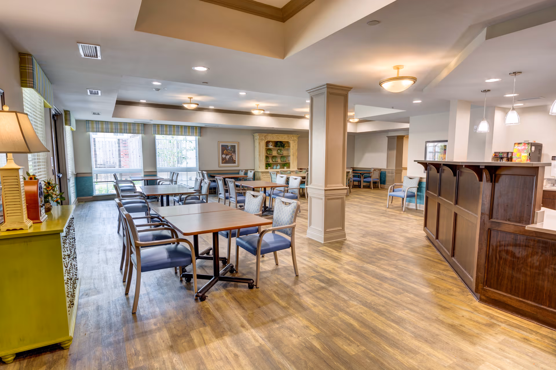 A spacious dining area with multiple tables and chairs arranged neatly. The room features large windows with striped valances allowing natural light to enter. There is a wooden counter on the right side with pendant lights hanging above it. The floor is wood-style, and the walls are painted in neutral tones with some decorative elements like a lamp on a green cabinet and framed artwork on the walls.