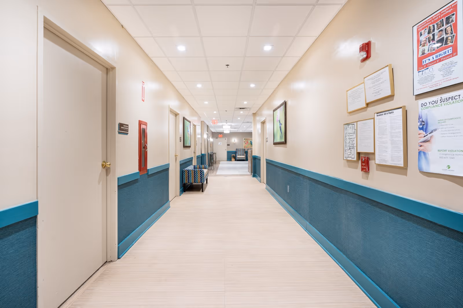 Well-lit indoor hallway of a senior care facility with beige walls, teal wainscoting, doors and seating along the corridor.