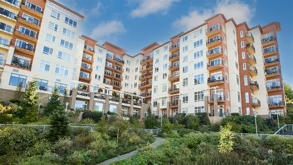 Exterior view of a multi-story senior living building with balconies and landscaped grounds under a blue sky.