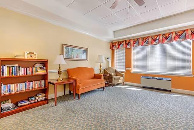 A cozy living room area with a patterned carpet, an orange loveseat, a wooden side table with a lamp, a wooden bookshelf filled with books, a wicker chair, and large windows with blinds and a colorful valance allowing natural light to fill the room.