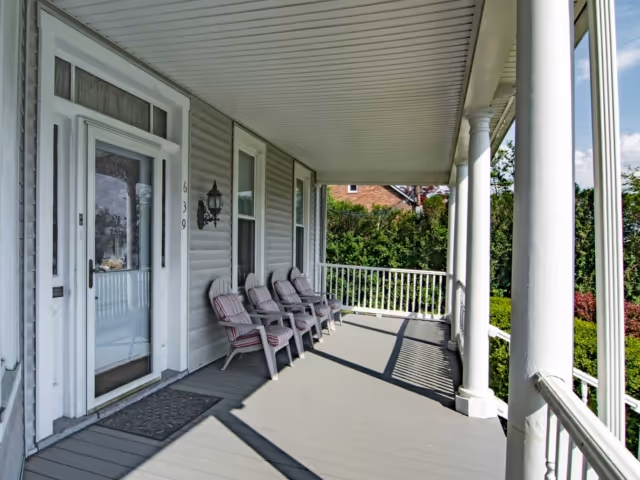 A covered porch area with four cushioned chairs lined up against the house wall. The porch has white railings and columns, with a gray floor and a white door with glass panels. Green bushes and trees are visible beyond the porch.