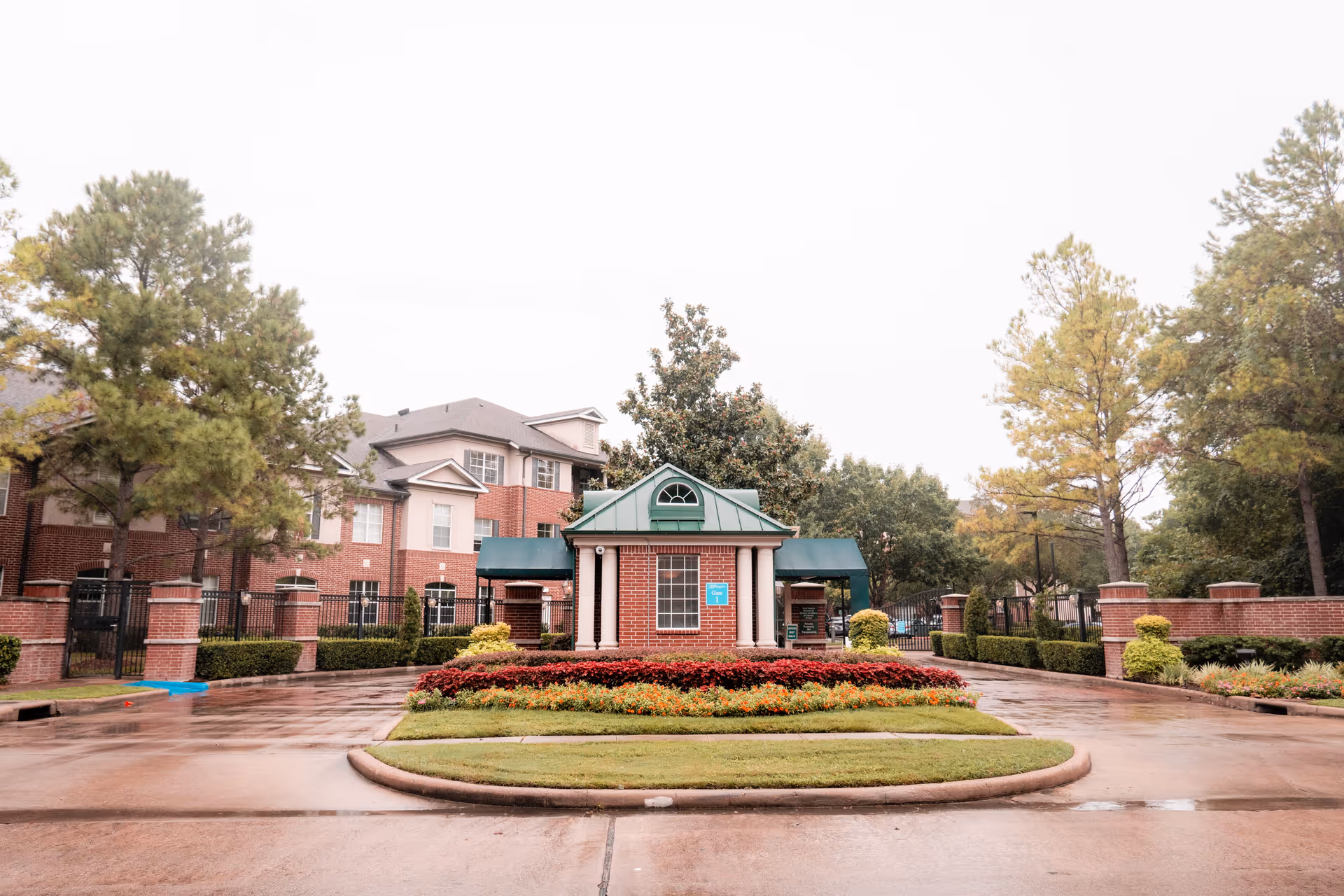 Entrance gatehouse of The Buckingham facility with a brick building, green roof, and surrounding landscaped flower beds and trees on a wet driveway.