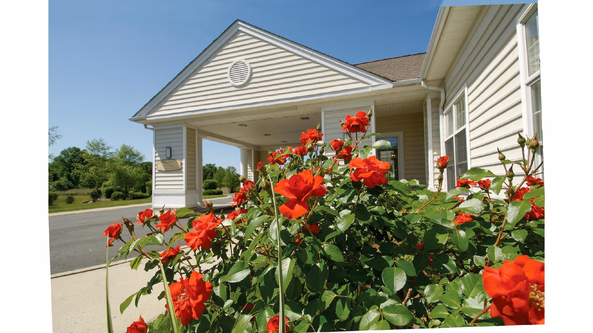 Entrance of a building with beige siding and a covered drop-off area, surrounded by vibrant red roses and greenery under a clear blue sky.