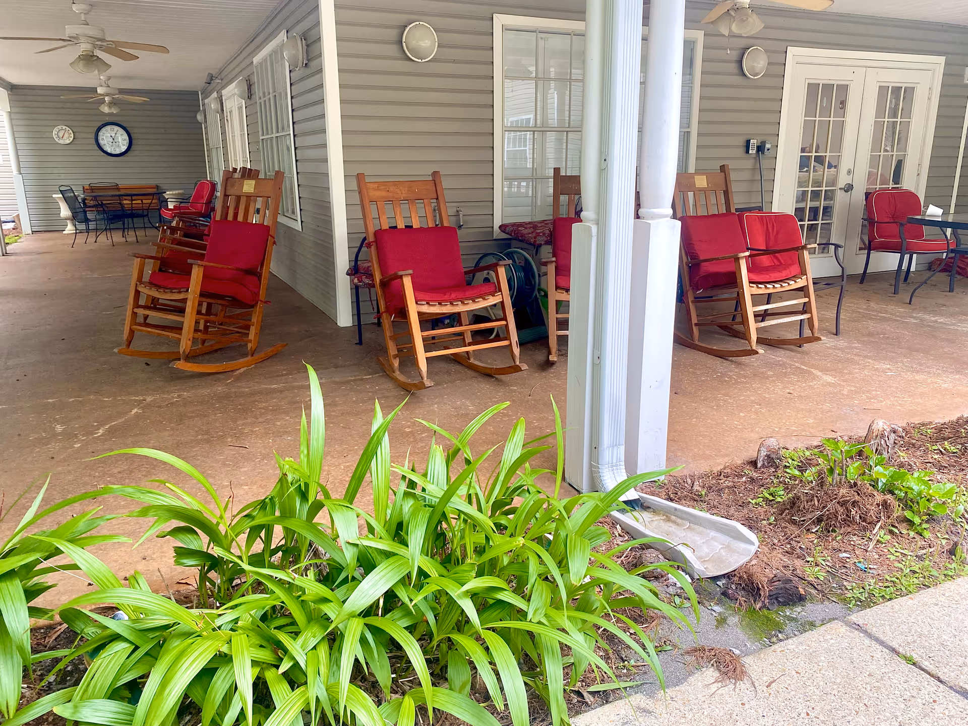 Covered outdoor patio area with several wooden rocking chairs with red cushions, ceiling fans, a table with chairs, and two clocks mounted on the wall. There are plants in the foreground and a downspout attached to a white column.